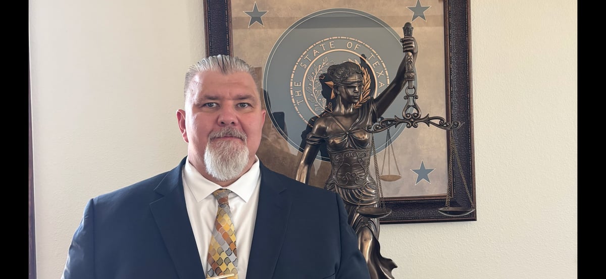 Man in dark suit and gold tie standing in front of an allegorical Lady Justice statue and the State of Texas seal