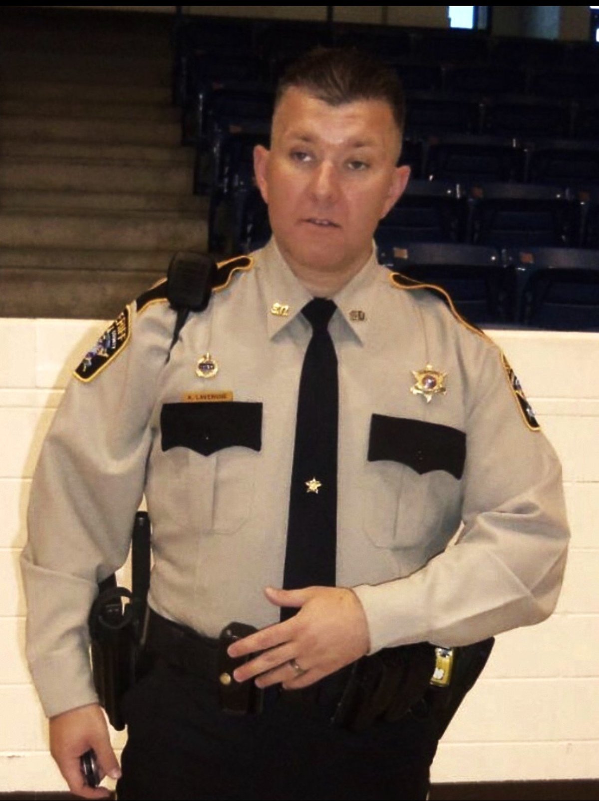 Law enforcement officer in tan and black uniform with badge and patches standing indoors