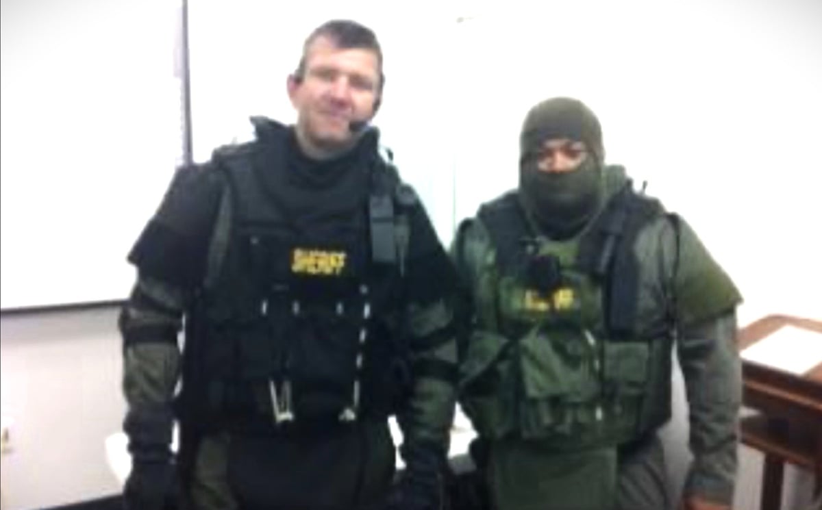 Two law enforcement officers in tactical gear and body armor standing indoors against a white wall