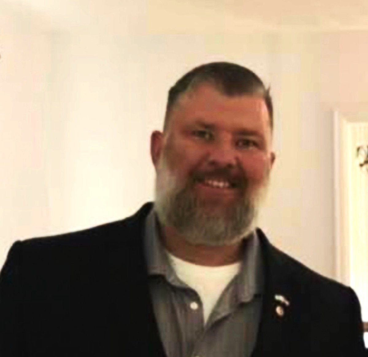 Portrait of a bearded man smiling at the camera wearing a black blazer and gray shirt indoors