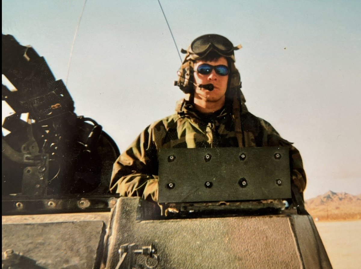 Military pilot in flight gear and sunglasses standing in aircraft cockpit over desert landscape